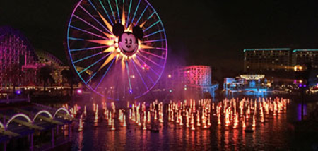 Ferris wheel with Mickey Mouse face brightly lit at night over water with fountains.