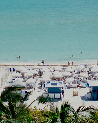Panoramic view of a crowded beach with turquoise water, white sand, colorful umbrellas, and palm trees.