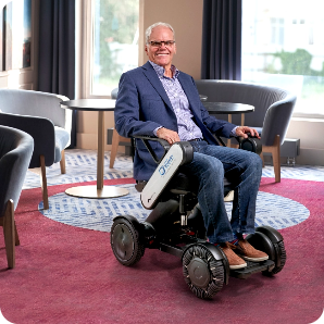 A smiling man in a blue blazer sits in a modern WHILL power wheelchair indoors.