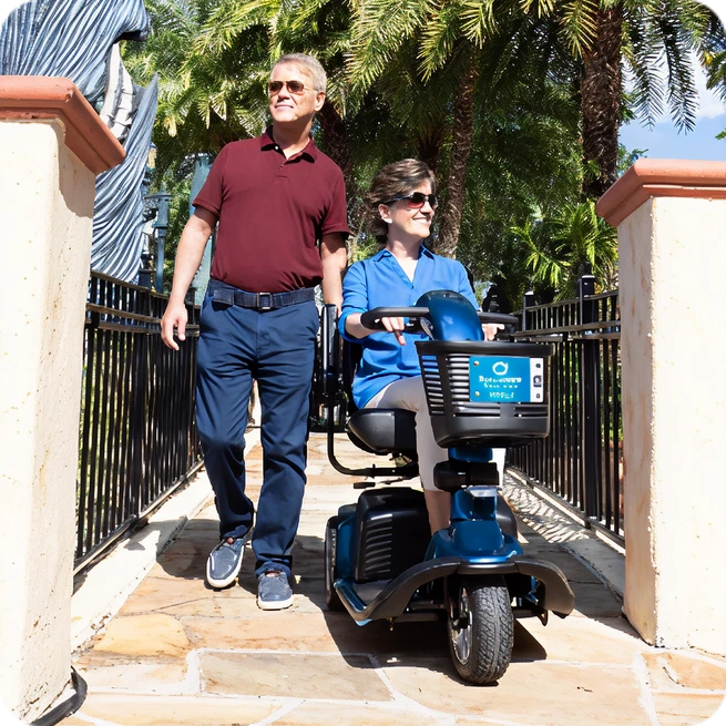 Smiling man walks next to woman riding a blue mobility scooter on a sunny pathway.
