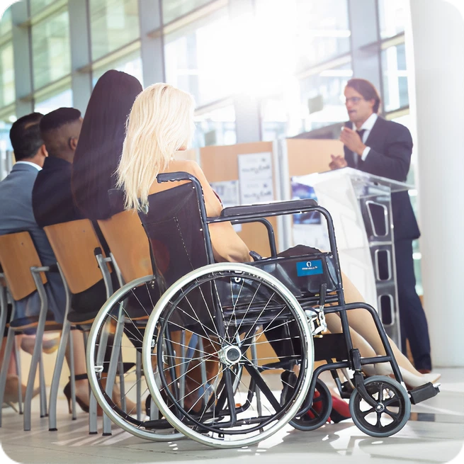 A woman in a wheelchair attends a professional conference, listening to a speaker.